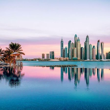 Dukes The Palm, A Royal Hideaway Hotel Dubai Exterior photo The image showcases a stunning skyline view of a modern city, likely during sunrise or sunset, as indicated by the beautiful pastel colors in the sky. The foreground features a calm body of water, reflecting the skyscrapers and the colorful sky. Lush