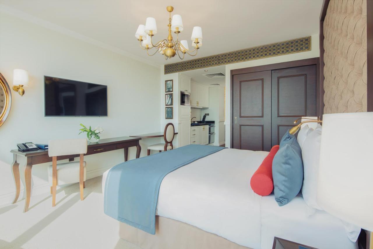 Dukes The Palm, A Royal Hideaway Hotel Dubai Exterior photo The photo depicts a modern hotel room featuring a neatly made queen-sized bed with decorative pillows in shades of blue and red. A stylish chandelier hangs from the ceiling, providing warm lighting. Opposite the bed, there is a wall-mounted flat-scre