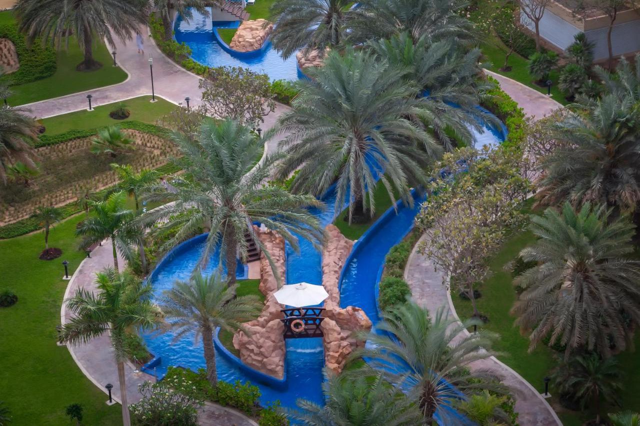 Dukes The Palm, A Royal Hideaway Hotel Dubai Exterior photo The photo shows a beautifully landscaped outdoor area featuring a winding water channel with vibrant blue water. Flanking the waterway are lush green palm trees and other vegetation, creating a tropical ambiance. There are also stone features along t