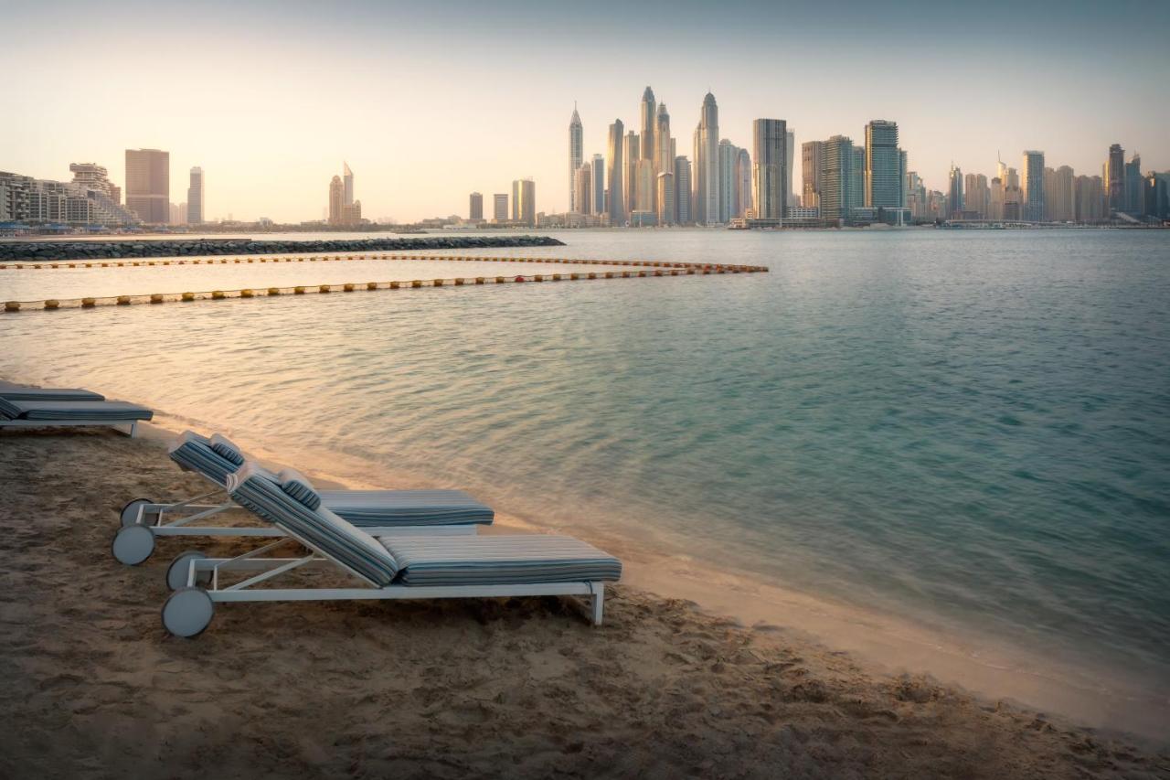Dukes The Palm, A Royal Hideaway Hotel Dubai Exterior photo The photo shows a tranquil beach scene with several lounge chairs lined up on the sand, facing a calm body of water. In the background, a modern skyline is visible, featuring tall buildings and skyscrapers. The atmosphere appears serene, possibly dur