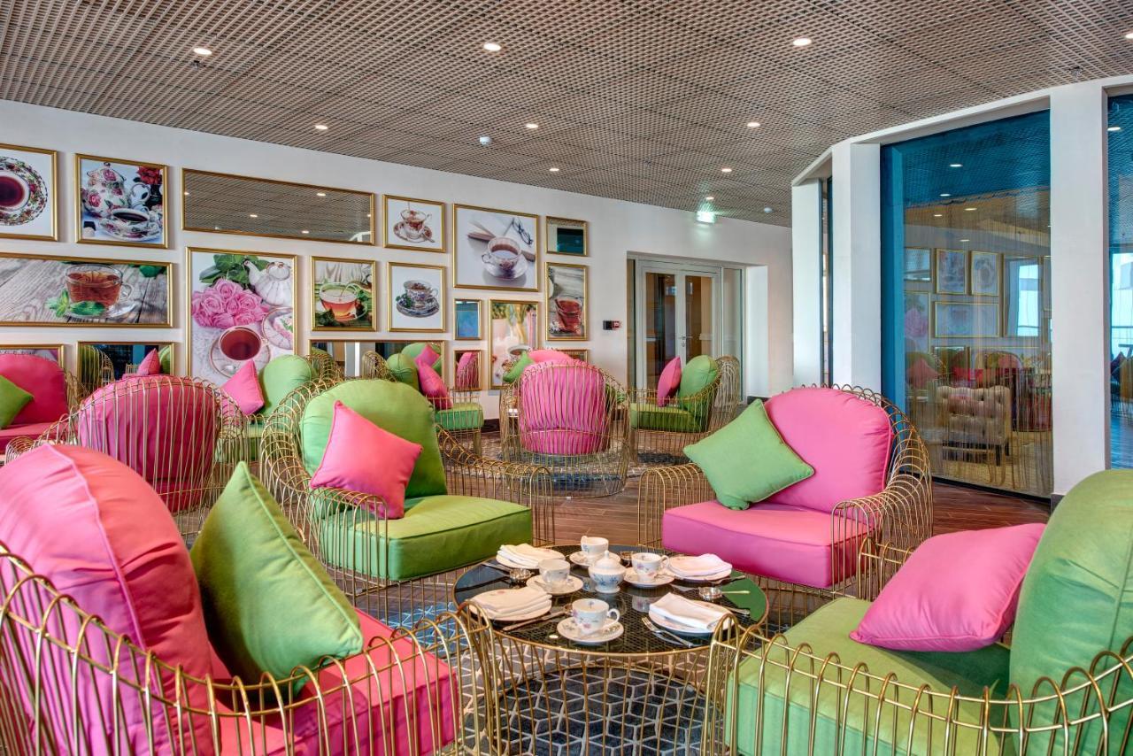 Dukes The Palm, A Royal Hideaway Hotel Dubai Exterior photo The photo shows a stylish and modern interior space, possibly a cafe or lounge. The seating area features elegant chairs with gold frames and vibrant cushions in shades of pink and green. There are several round tables with tea sets and small plates 