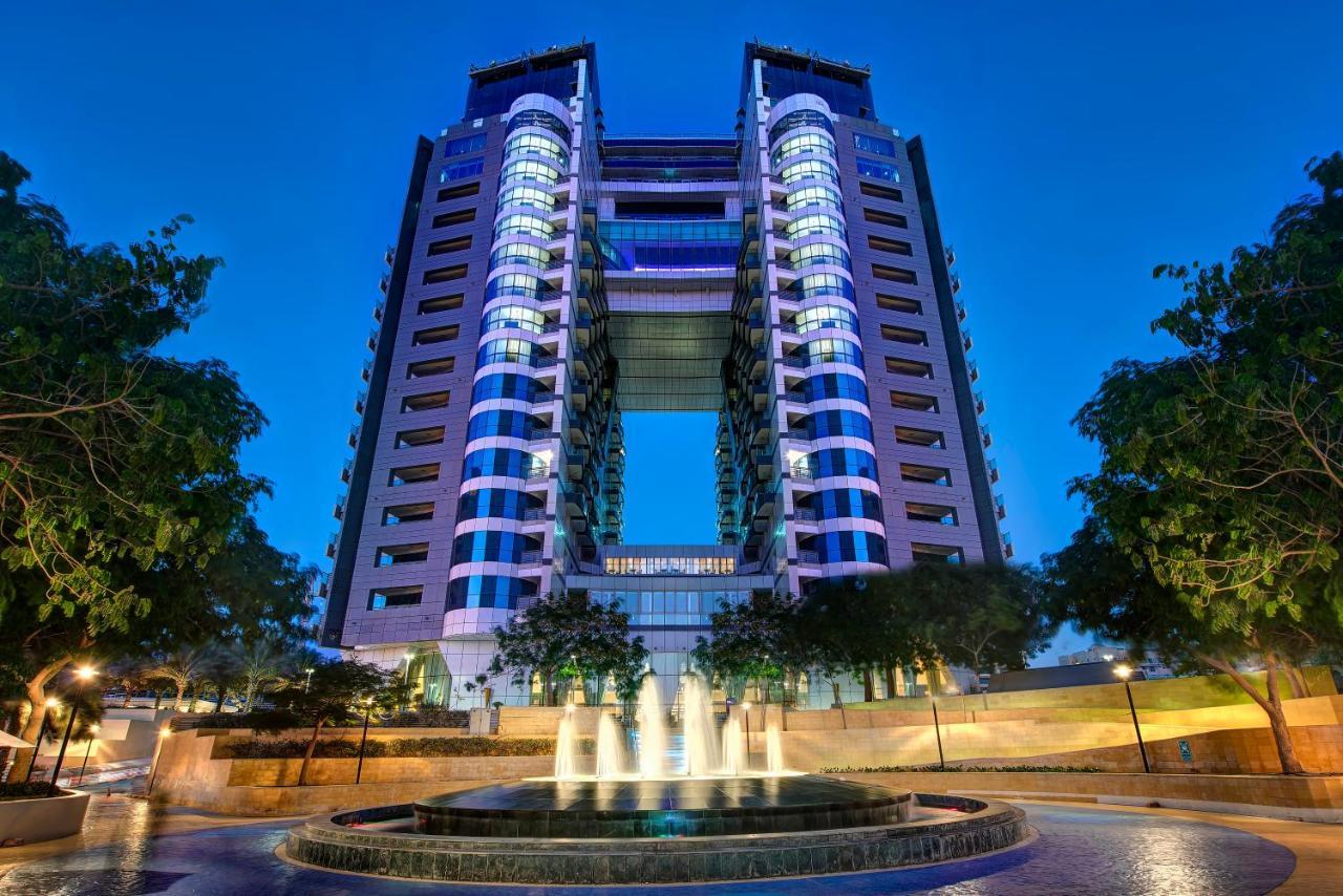 Dukes The Palm, A Royal Hideaway Hotel Dubai Exterior photo The photo shows a modern, tall building with a distinctive architectural design, featuring multiple floors and large windows. The structure is illuminated at dusk, highlighting its design and colors. In the foreground, there is a circular fountain wi