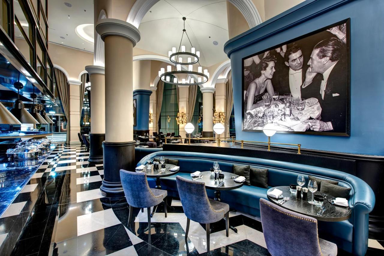 Dukes The Palm, A Royal Hideaway Hotel Dubai Exterior photo The image shows an elegant restaurant interior with a sophisticated design. The space features high ceilings, large columns, and a combination of blue and black color schemes. There is a stylish seating area with upholstered chairs and tables set for