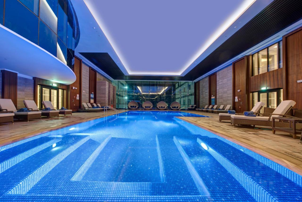 Dukes The Palm, A Royal Hideaway Hotel Dubai Exterior photo The photo shows a luxurious indoor swimming pool area. The pool is rectangular and features a blue tiled bottom, creating a vibrant hue in the water. Surrounding the pool are comfortable lounge chairs lined up along the edges. The area has large glas