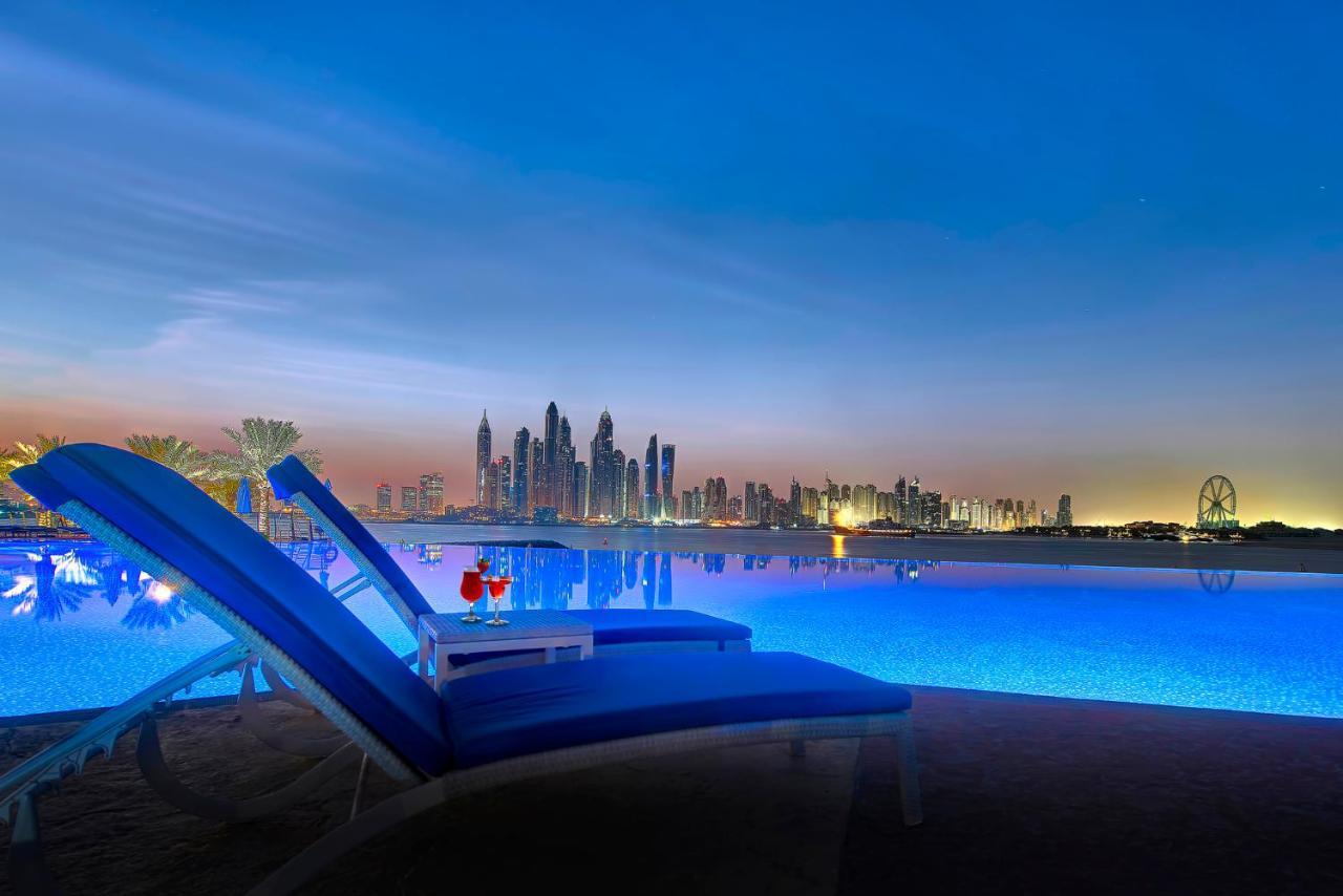 Dukes The Palm, A Royal Hideaway Hotel Dubai Exterior photo The image depicts a tranquil scene by a swimming pool, illuminated by soft blue lighting. In the foreground, there are two lounge chairs positioned beside the pool, suggesting a relaxing environment. A small table between the chairs holds two cocktai