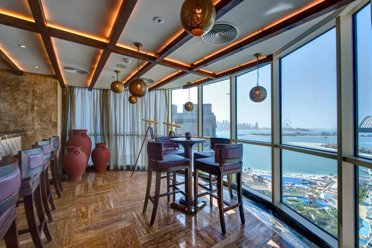 Dukes The Palm, A Royal Hideaway Hotel Dubai Exterior photo The photo depicts a modern, stylish interior of a restaurant or lounge. It features large windows that offer a view of a waterfront and cityscape. The design includes warm wooden beams and ambient lighting, creating a cozy atmosphere. There are sever