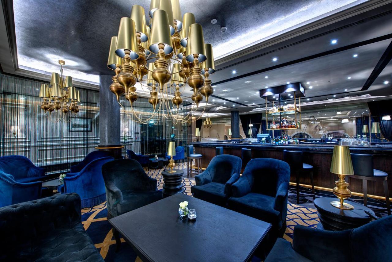 Dukes The Palm, A Royal Hideaway Hotel Dubai Exterior photo The photo displays an elegant interior of a lounge or bar area. It features plush blue seating, including armchairs around a dark table. A prominent chandelier with golden lamps hangs from the ceiling, providing ambient light. In the background, ther