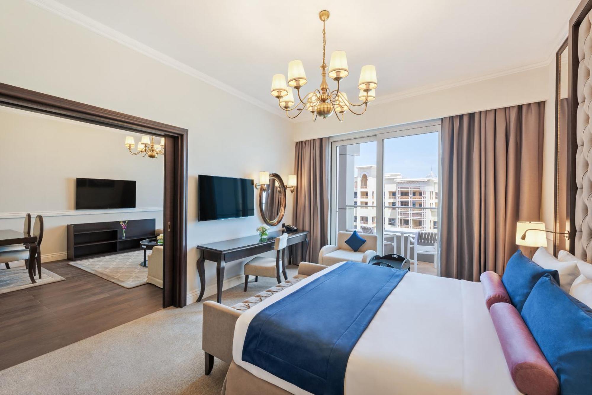 Dukes The Palm, A Royal Hideaway Hotel Dubai Exterior photo The photo depicts a stylish hotel room. It features a comfortable bed with blue and white bedding. The room includes a large window with curtains that allow natural light in, and there are two flat-screen TVs mounted on the walls. A chandelier hangs 