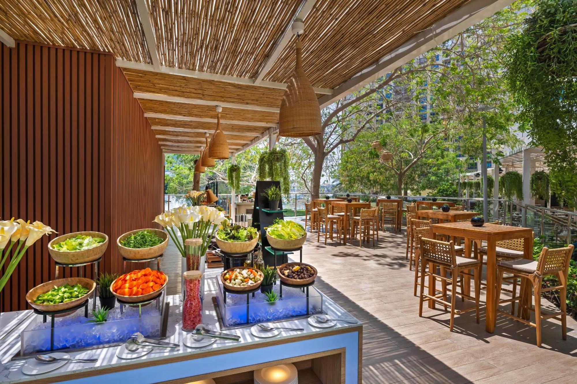 Dukes The Palm, A Royal Hideaway Hotel Dubai Exterior photo The photo showcases a bright and inviting outdoor dining space. In the foreground, there is a buffet table with various salads and fresh vegetables displayed in bowls, including items like lettuce, carrots, and other colorful ingredients, set against