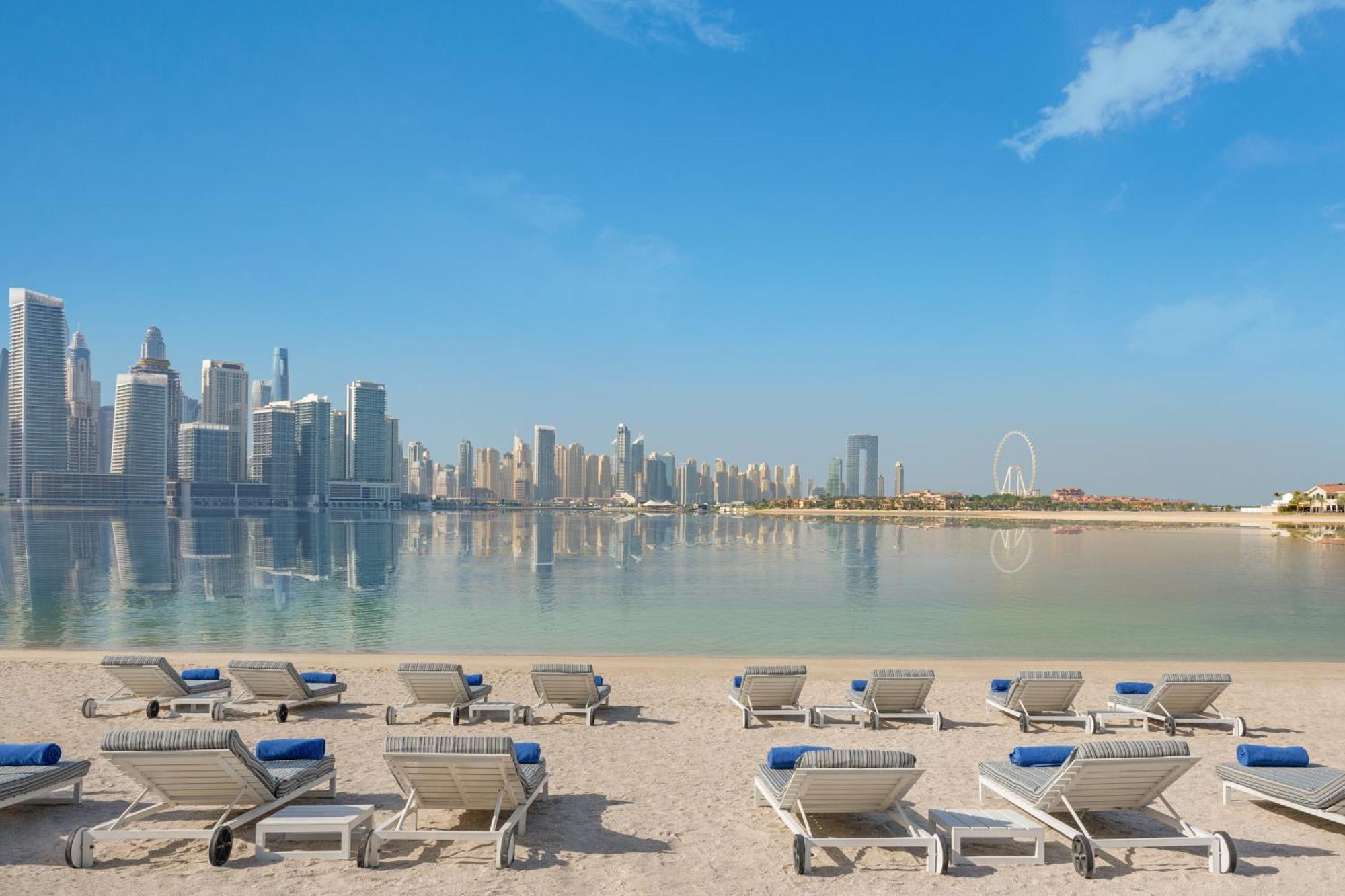 Dukes The Palm, A Royal Hideaway Hotel Dubai Exterior photo The photo shows a serene beach scene with several lounge chairs arranged in the foreground. These chairs are positioned on soft sand and have blue cushions. In the background, there is a stunning view of a modern skyline featuring tall buildings and 