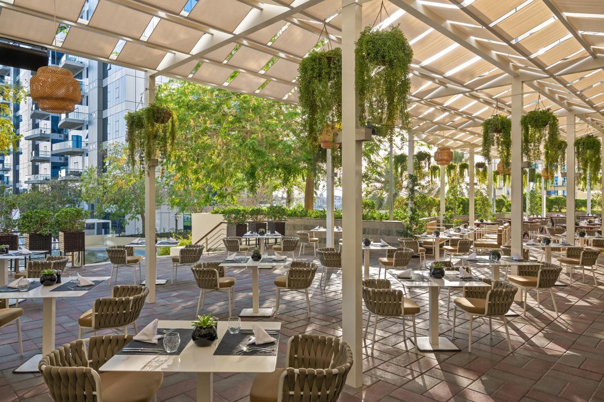 Dukes The Palm, A Royal Hideaway Hotel Dubai Exterior photo The photo shows a charming outdoor restaurant or café with a spacious seating area. The space is adorned with several plants hanging from the ceiling, creating a vibrant and lush atmosphere. There are rows of tables set with utensils and small decora