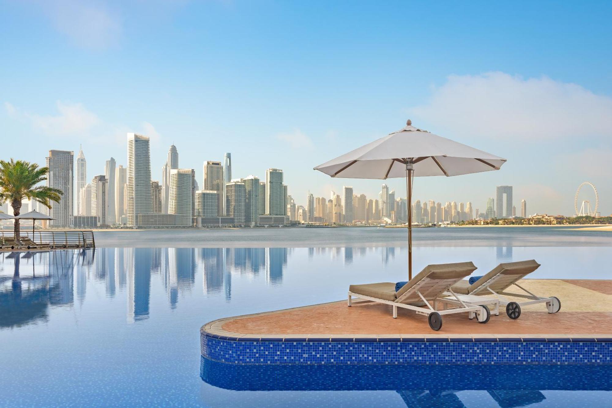 Dukes The Palm, A Royal Hideaway Hotel Dubai Exterior photo The photo shows a serene and luxurious setting, featuring an infinity pool that seems to blend into a body of water. In the foreground, there are two lounge chairs positioned on a platform, accompanied by a large white umbrella providing shade. The b