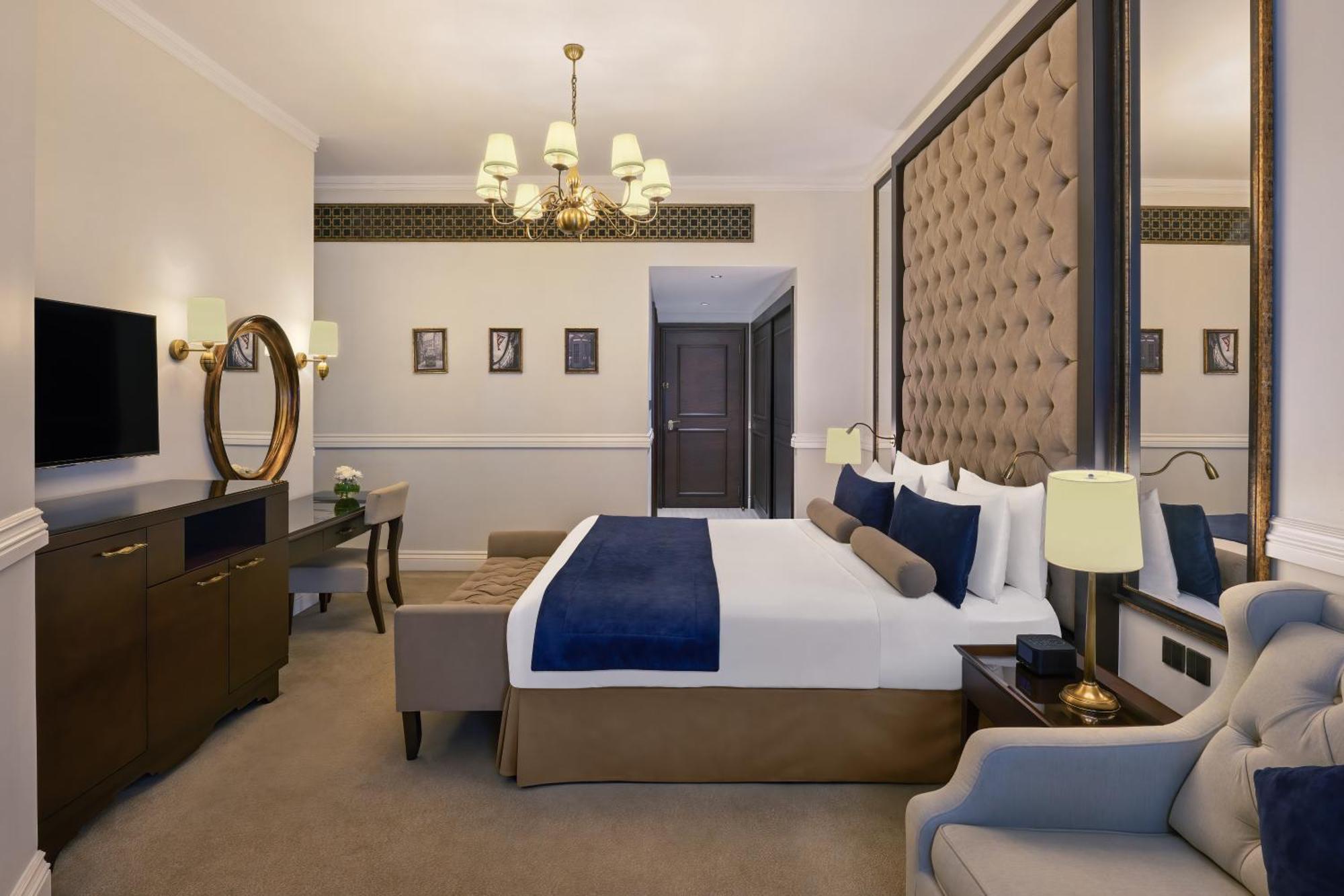 Dukes The Palm, A Royal Hideaway Hotel Dubai Exterior photo The photo shows a stylish hotel room. It features a large bed with white linens and a blue throw blanket, which is placed against a luxurious padded wall. On either side of the bed, there are bedside lamps. A modern chandelier hangs from the ceiling,