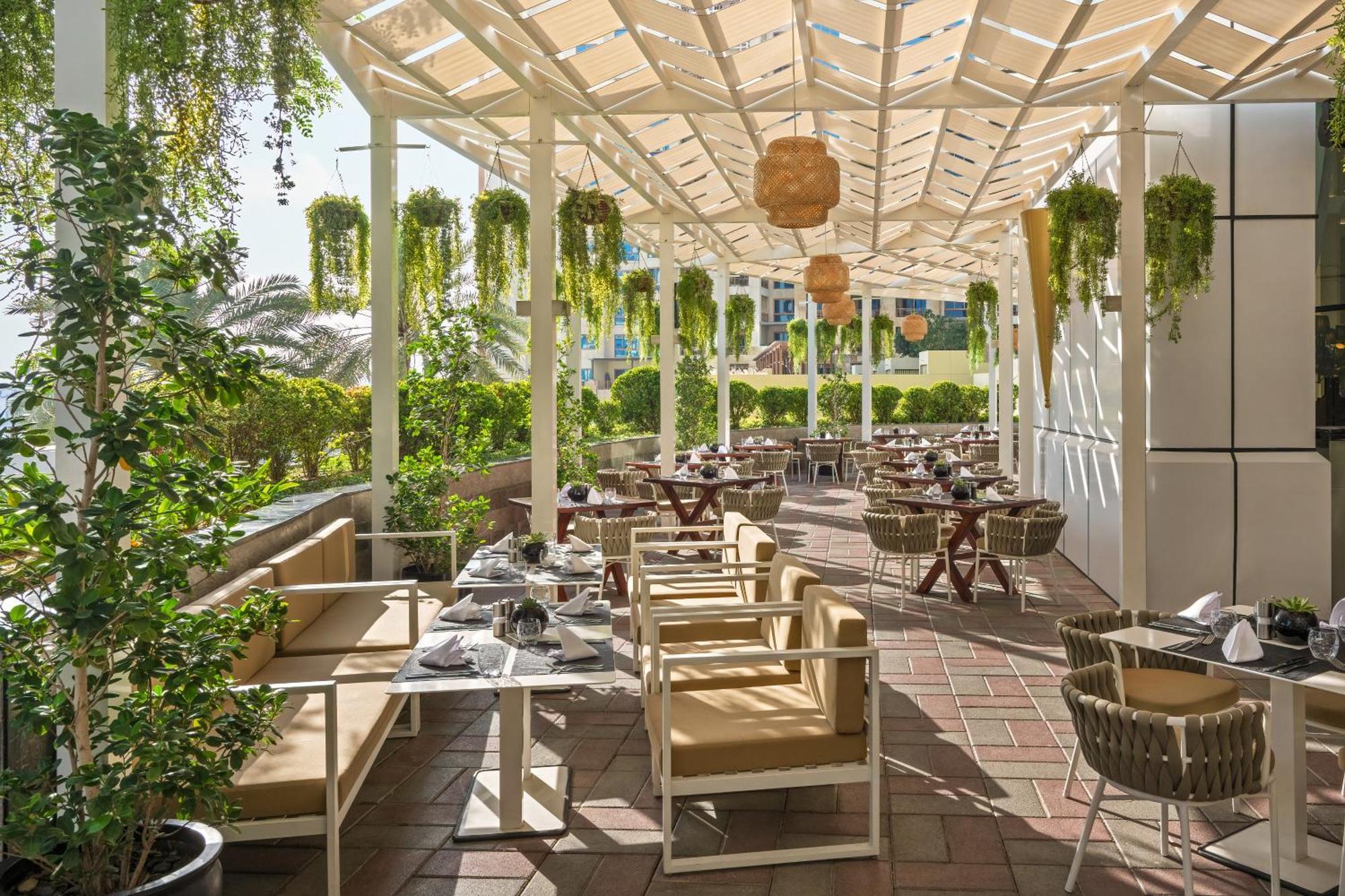 Dukes The Palm, A Royal Hideaway Hotel Dubai Exterior photo The photo shows an outdoor restaurant patio area with a bright and inviting atmosphere. There are several tables and chairs arranged neatly under a structured canopy. The furniture appears to be a mix of light-colored wooden pieces and wicker chairs.