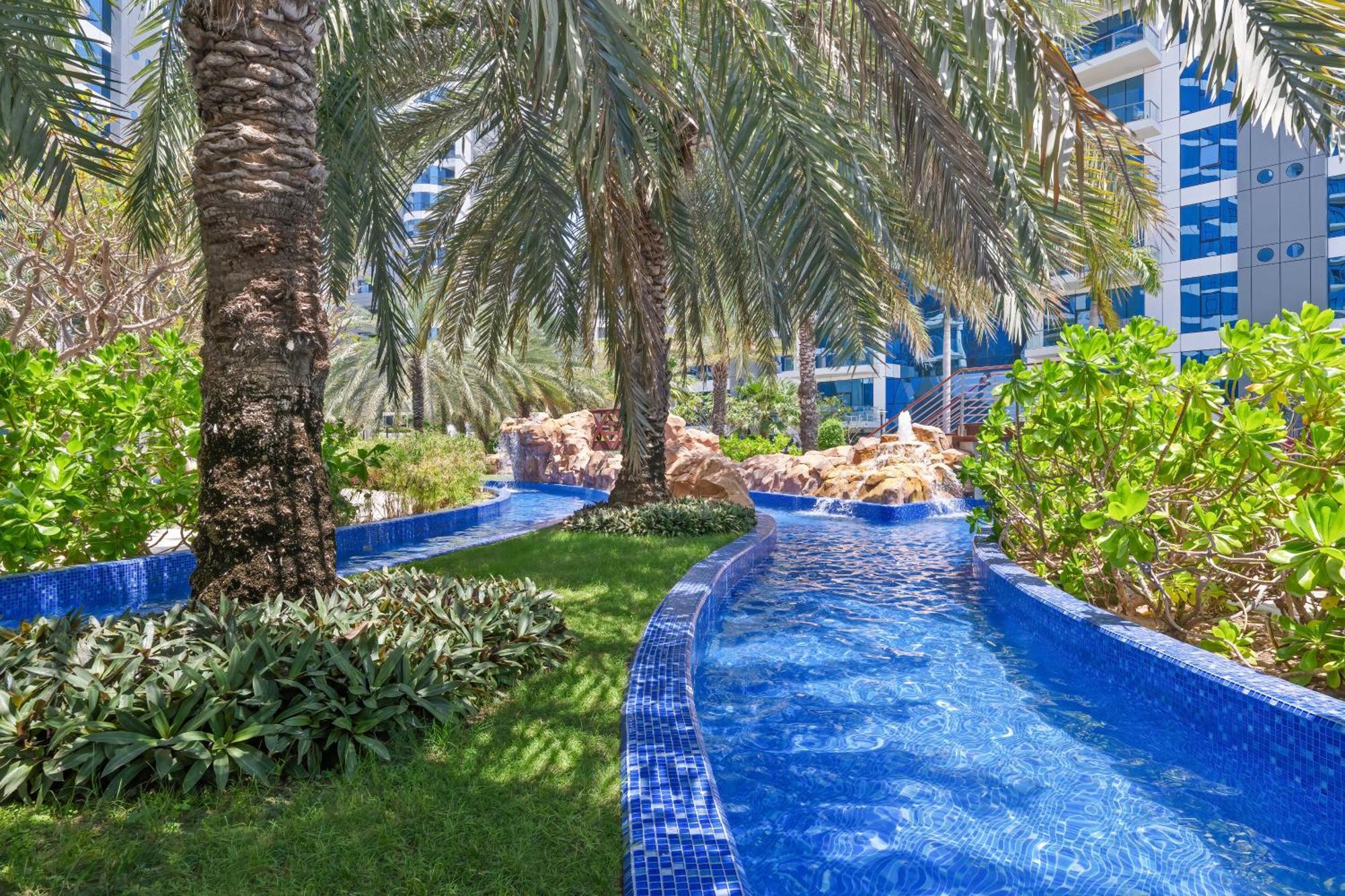 Dukes The Palm, A Royal Hideaway Hotel Dubai Exterior photo The photo depicts a beautifully landscaped tropical area featuring a winding waterway with clear blue water, bordered by lush greenery and palm trees. The vibrant blue tiles lining the water add a decorative touch, while the overall setting suggests 