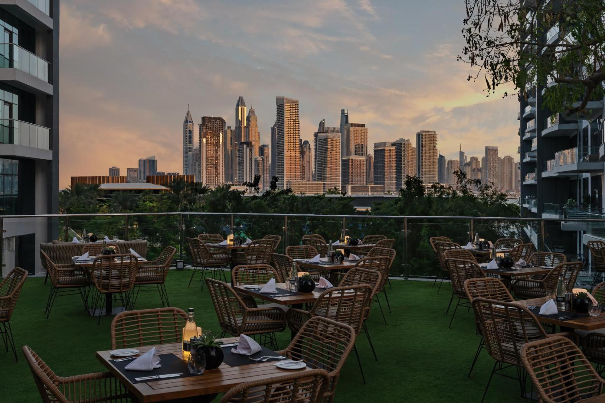 Dukes The Palm, A Royal Hideaway Hotel Dubai Exterior photo The photo shows an outdoor dining area with several tables and chairs arranged neatly. The tables are set with plates and candles, creating an inviting atmosphere. In the background, there is a stunning city skyline featuring tall skyscrapers, bathed