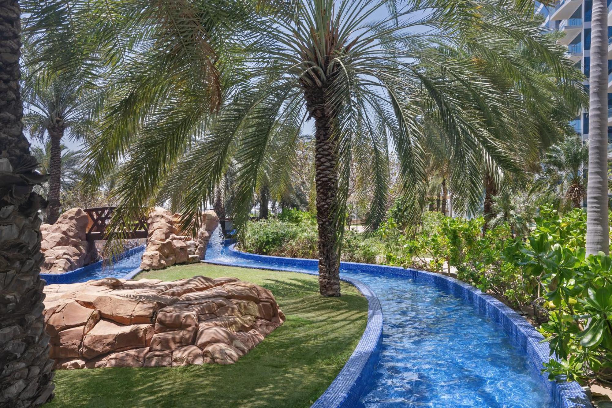 Dukes The Palm, A Royal Hideaway Hotel Dubai Exterior photo The photo depicts a serene outdoor setting, featuring a winding blue water channel surrounded by lush greenery. Palm trees tower over the scene, providing shade and a tropical feel. There are rocky formations integrated into the landscape, and the ov