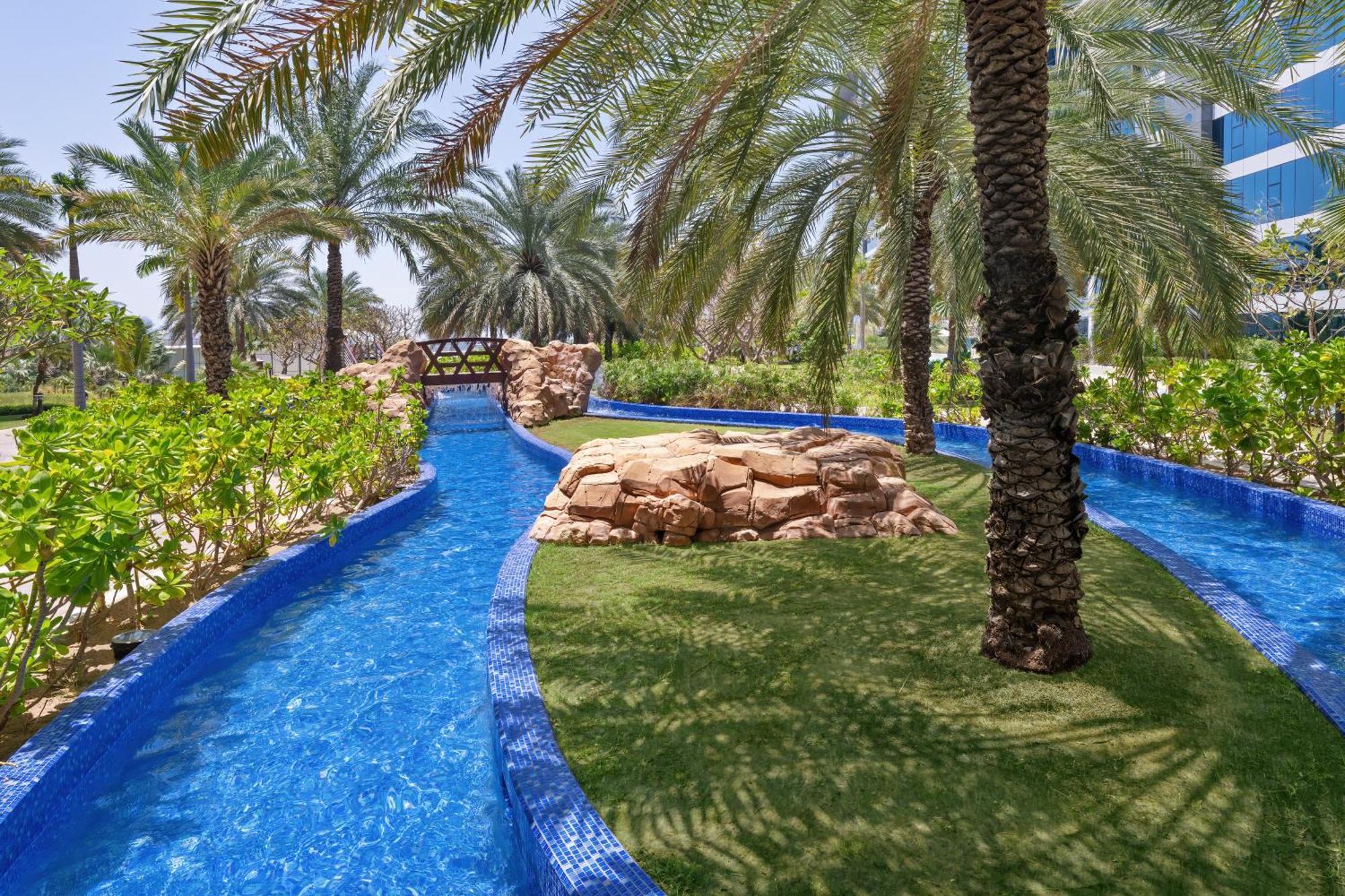 Dukes The Palm, A Royal Hideaway Hotel Dubai Exterior photo The photo depicts a serene outdoor area featuring a winding, clear blue waterway surrounded by lush greenery and palm trees. In the background, there is a small stone bridge arching over the water. Natural rock formations and manicured grass create a