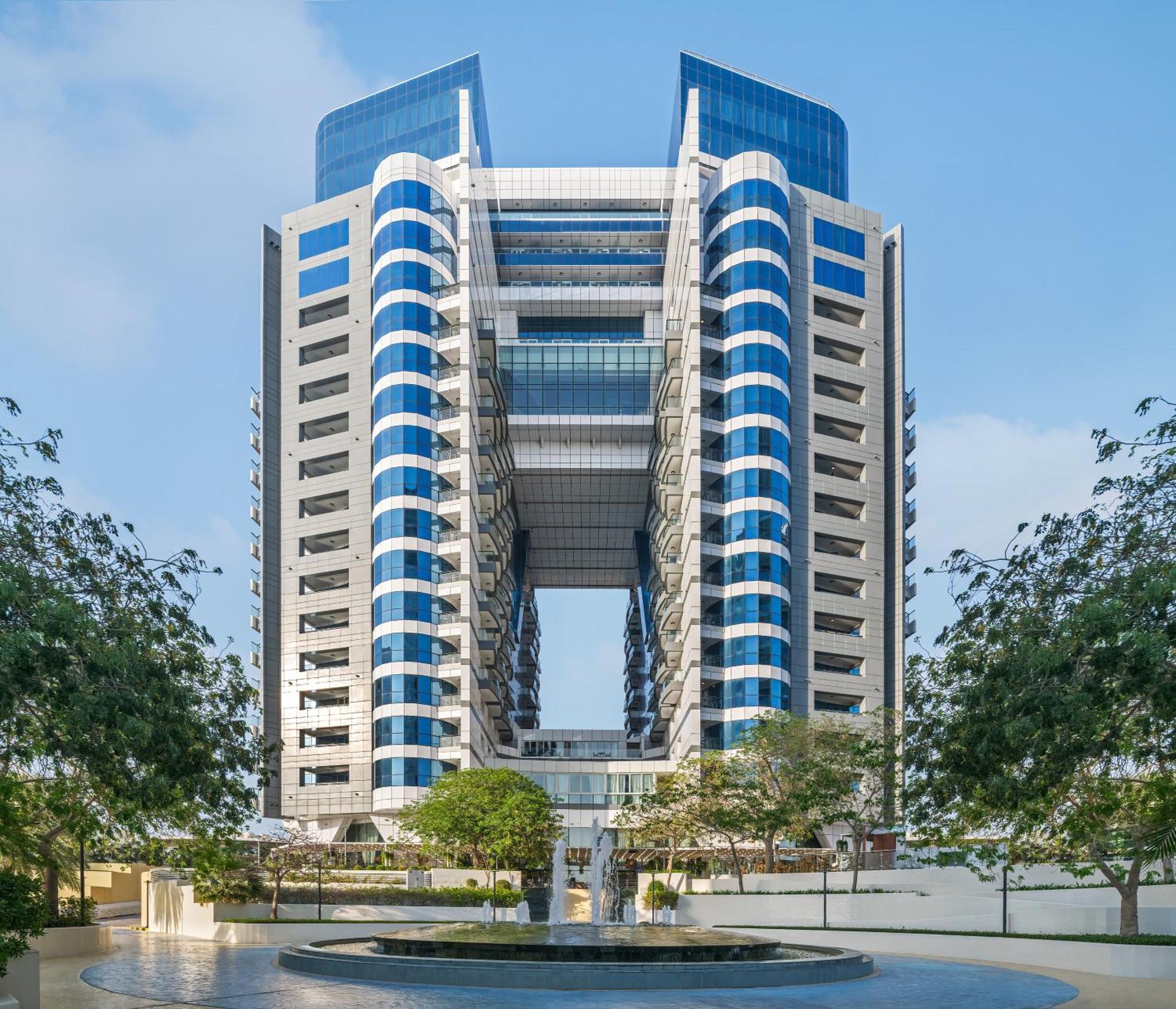 Dukes The Palm, A Royal Hideaway Hotel Dubai Exterior photo The image shows a large modern building characterized by a distinctive architectural design. It features a symmetrical façade with multiple levels and large, blue-tinted glass windows. The structure has prominent vertical elements and an open center,
